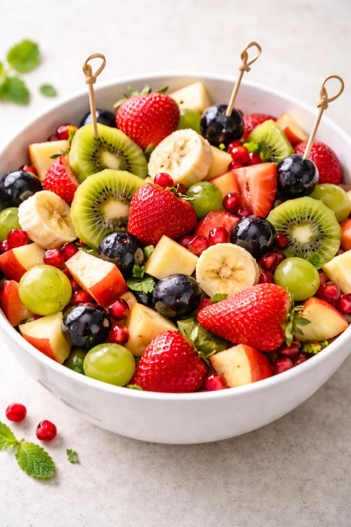 Fresh mixed fruit salad with kiwi, strawberries, banana, grapes, apple, and pomegranate seeds served in white ceramic bowl close up view